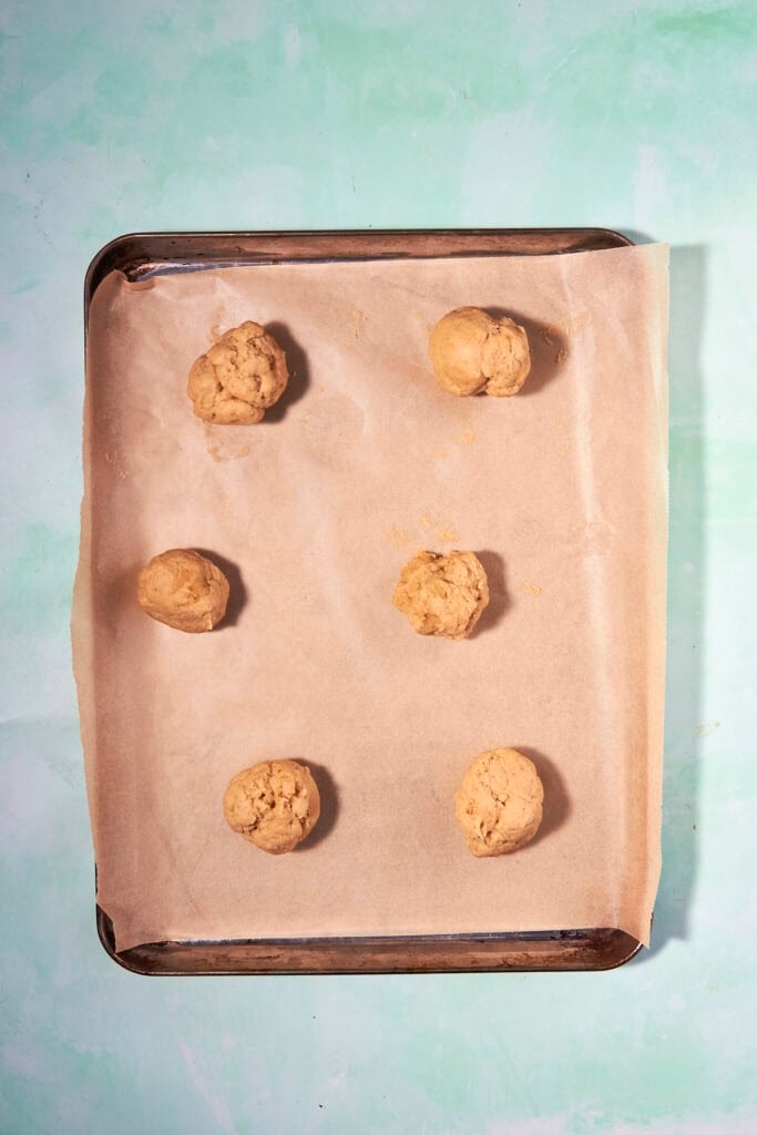 Six unbaked banana bread cookies sit spaced apart on a parchment-lined baking sheet, set against a light turquoise background.