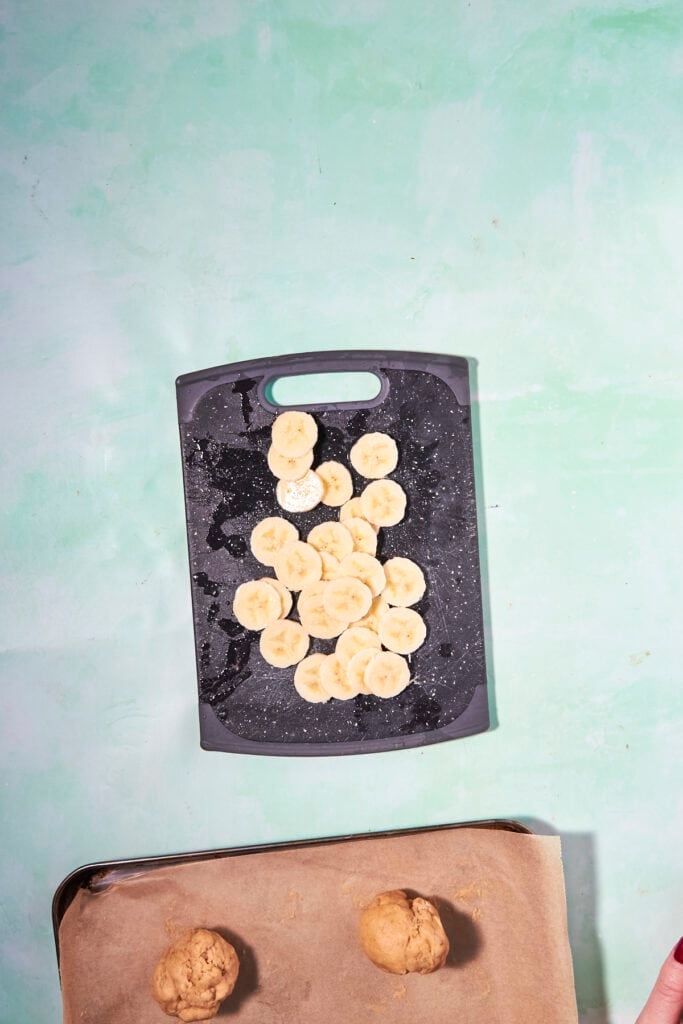 A black cutting board with sliced bananas sits on a light green surface. Below it, a baking sheet lined with parchment holds two balls of banana bread cookie dough.