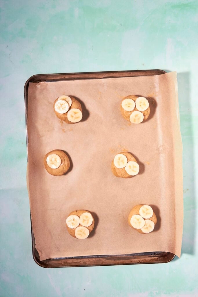 A baking sheet lined with parchment paper holds six unbaked banana bread cookies, each dough ball topped with banana slices, against a light green background.