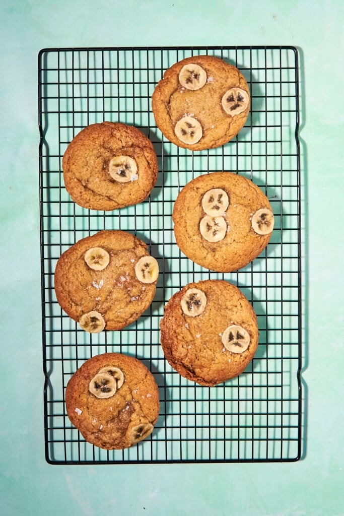 Six large banana bread cookies topped with banana slices are arranged on a black wire cooling rack against a light blue background.