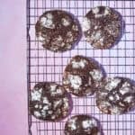 Six chocolate crinkle cookies dusted with powdered sugar are cooling on a wire rack, set against a pink background.