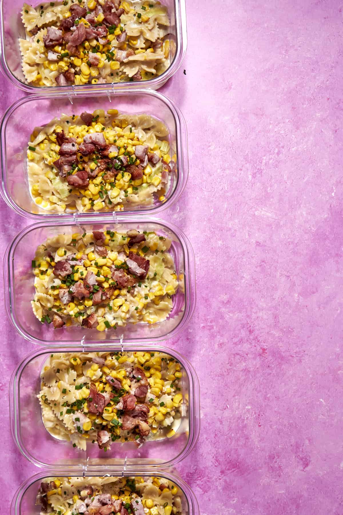 Five glass meal prep containers filled with bowtie pasta, roasted corn, and diced ham are arranged in a vertical row on a bright pink background. The containers are neatly organized, with food garnished with herbs.