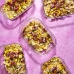 Five clear containers filled with bow-tie pasta, roasted corn, chives, and bacon pieces sit on a bright pink surface. The pasta is evenly distributed, and the toppings add color and texture to each portion.