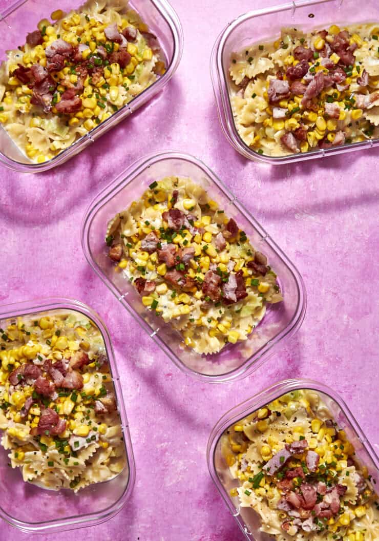 Five clear containers filled with bow-tie pasta, roasted corn, chives, and bacon pieces sit on a bright pink surface. The pasta is evenly distributed, and the toppings add color and texture to each portion.