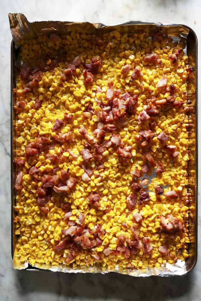 A baking sheet lined with foil, covered in roasted corn and crumbled cooked bacon pieces on a marble surface.