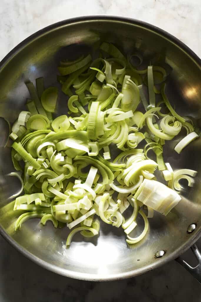 Sliced leeks in a stainless steel pan, ready to be cooked, with light shining on them from the side—perfect for pairing with roasted corn for a delicious meal.