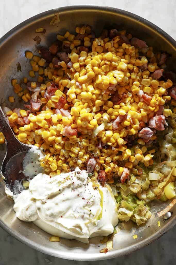 A metal pan filled with roasted corn, diced ham, sautéed onions, and a large dollop of creamy white sauce, all being mixed with a wooden spoon.