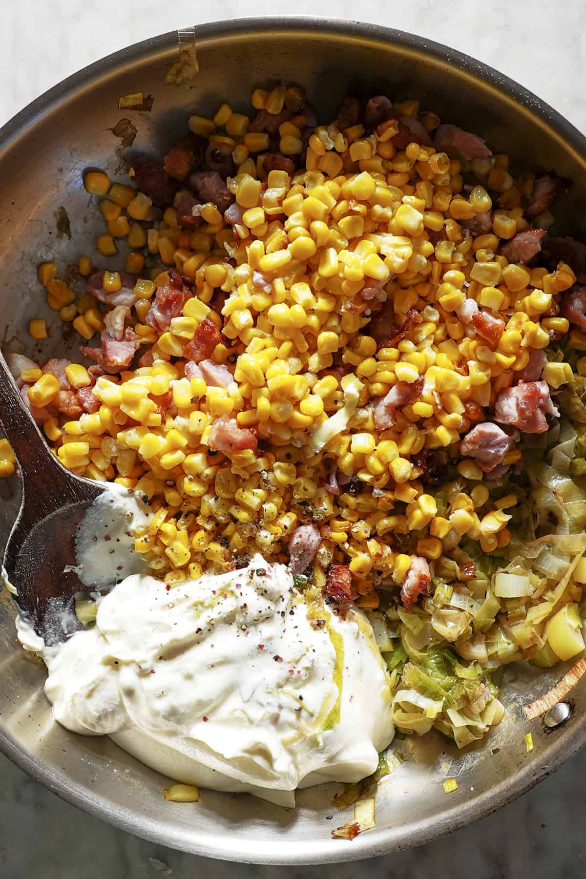A metal mixing bowl filled with roasted corn, diced ham, cooked onions, and a generous dollop of sour cream being mixed with a wooden spoon on a marble countertop.