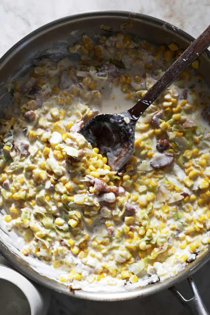A skillet filled with creamy roasted corn, bacon, and chopped green peppers being stirred with a wooden spoon on a marble countertop.