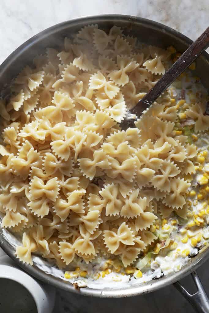 A close-up of a skillet containing cooked farfalle pasta mixed with creamy sauce, roasted corn, and vegetables, being stirred with a wooden spoon on a marble surface.