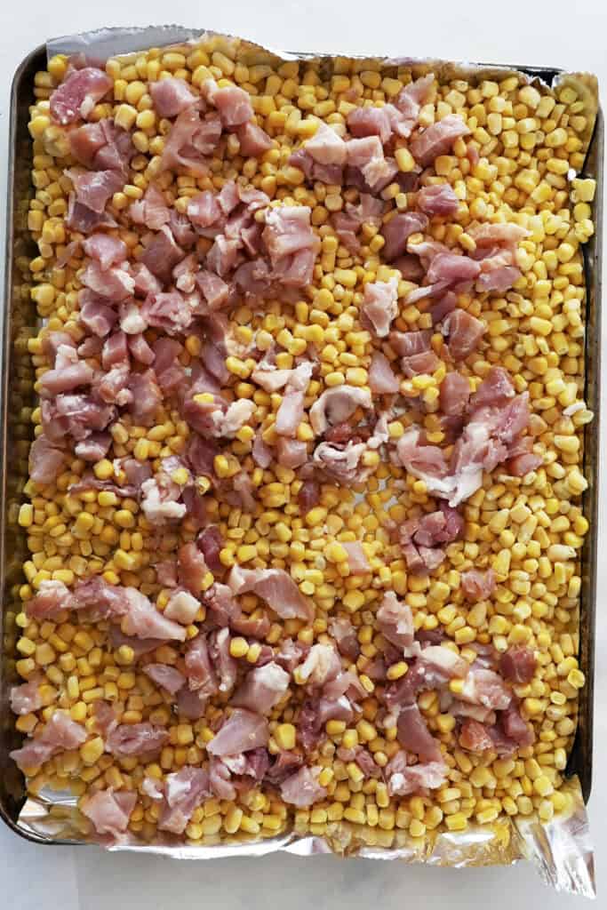 A baking sheet lined with foil, covered with a layer of roasted corn kernels and topped with evenly distributed pieces of raw, diced chicken.