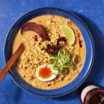 A bowl of ramen with creamy broth reminiscent of Buldak Soup, topped with a halved soft-boiled egg, sliced scallions, chili flakes, and a lime wedge. A wooden spoon rests in the bowl, and a jar of chili oil sits nearby on a blue surface.