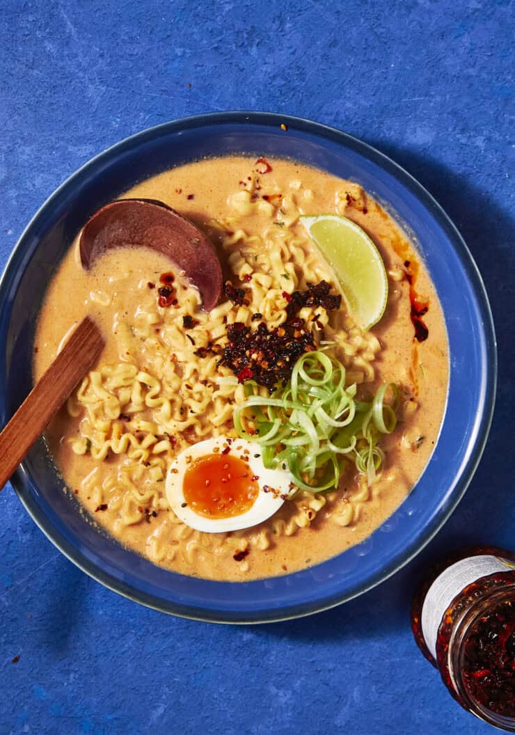 A bowl of ramen with creamy broth reminiscent of Buldak Soup, topped with a halved soft-boiled egg, sliced scallions, chili flakes, and a lime wedge. A wooden spoon rests in the bowl, and a jar of chili oil sits nearby on a blue surface.