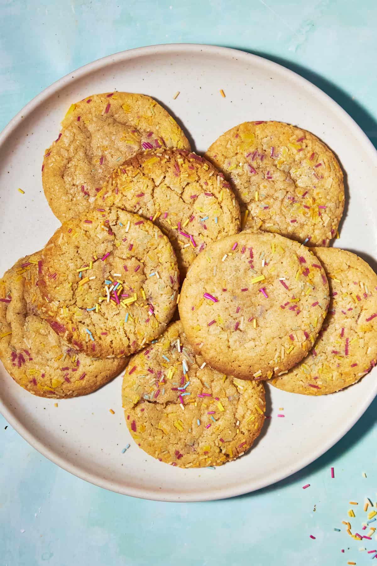 A plate of seven golden brown cookies topped with colorful sprinkles, arranged on a white plate against a light turquoise background.