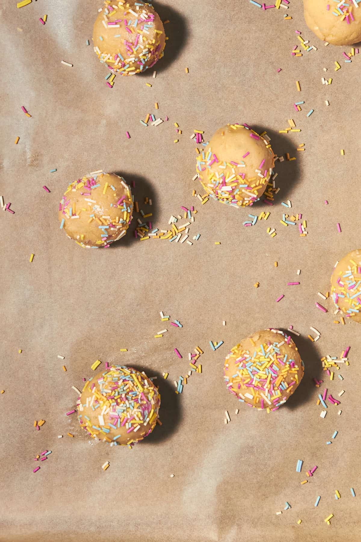 Cookie dough balls topped with colorful sprinkles are arranged on a sheet of parchment paper, with extra sprinkles scattered around them.
