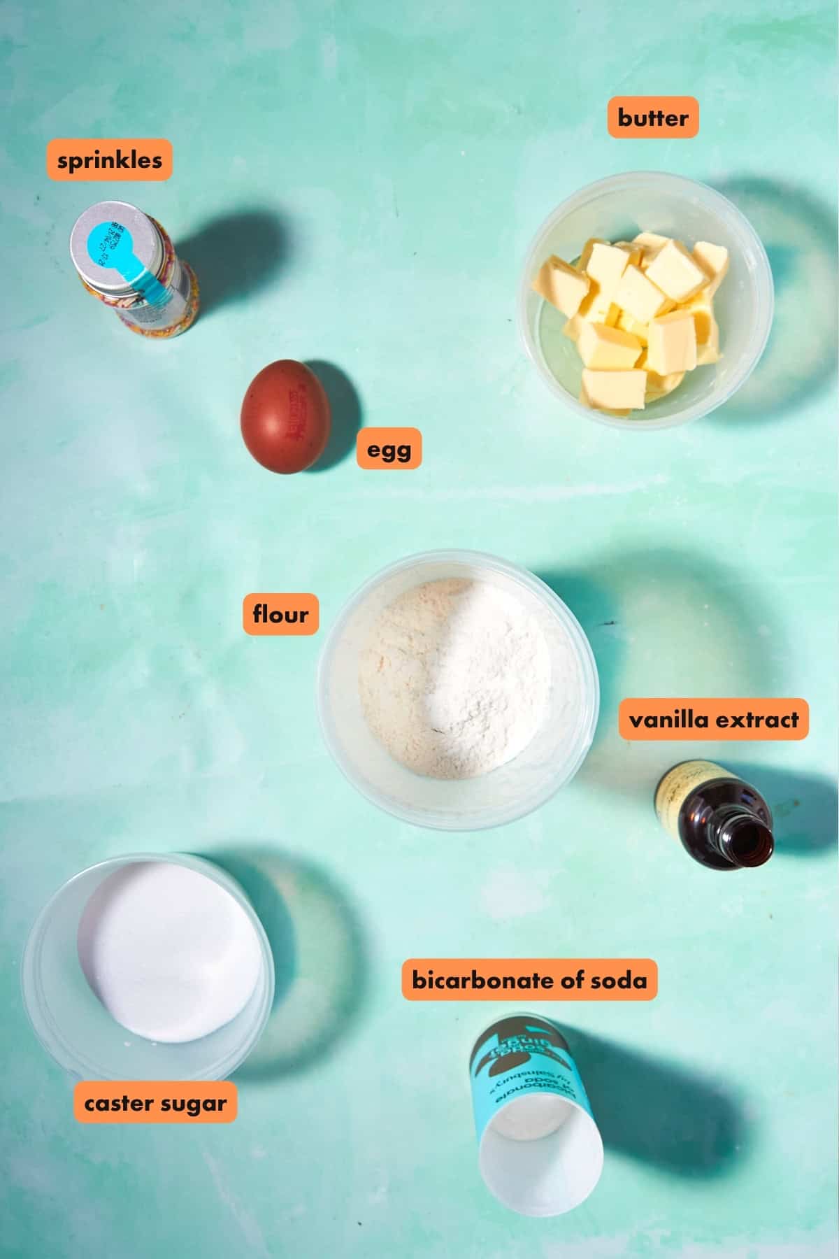 A flat lay of baking ingredients on a turquoise surface for Funfetti Cookies: sprinkles, a brown egg, cubed butter, flour, vanilla extract, caster sugar, and bicarbonate of soda. Each item is labeled with orange tags.