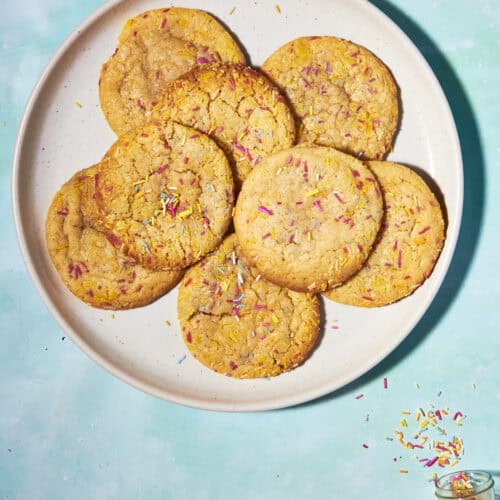 A plate of eight golden cookies with colorful sprinkles sits on a light blue surface, with extra sprinkles scattered nearby and spilling from a small jar.