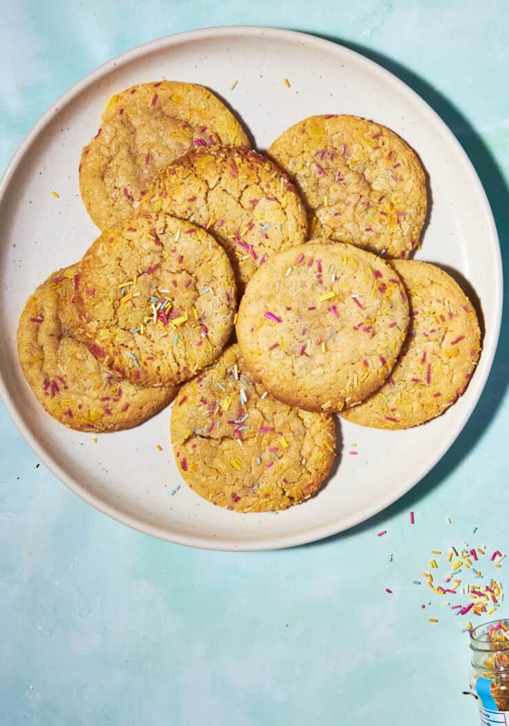 A plate of eight golden cookies with colorful sprinkles sits on a light blue surface, with extra sprinkles scattered nearby and spilling from a small jar.