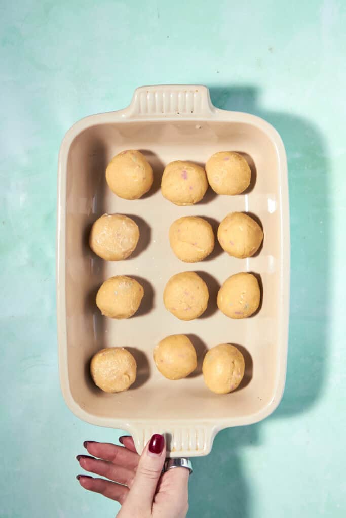 A hand with red nail polish holds a beige baking dish containing twelve evenly spaced raw dough balls on a turquoise background.