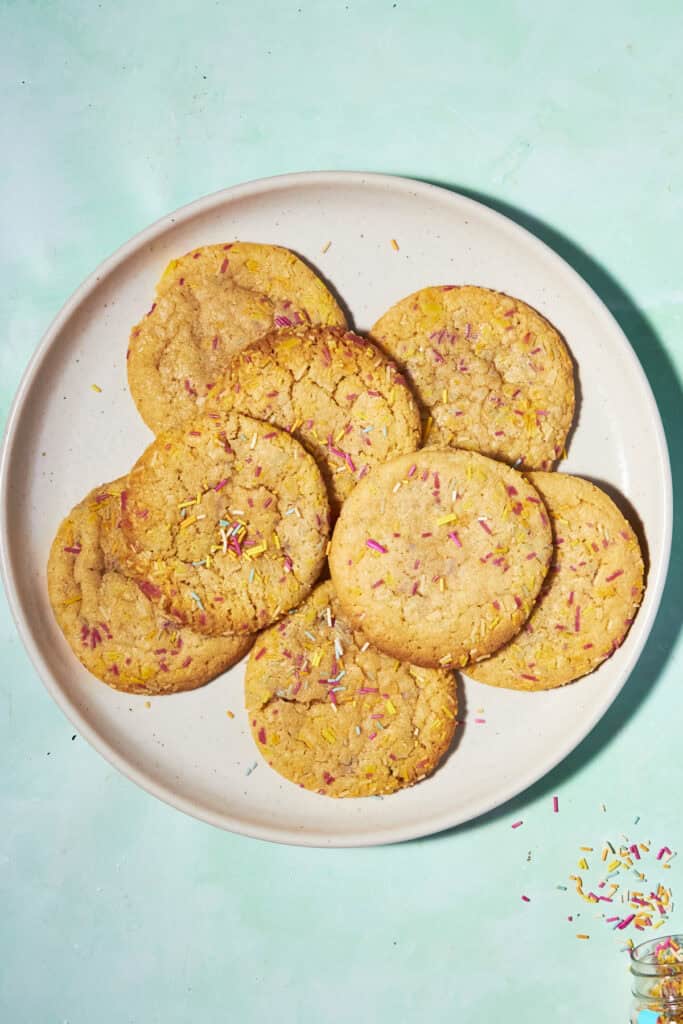 A plate of eight golden brown cookies topped with colorful sprinkles, arranged in a circular pattern on a light-colored plate against a pastel turquoise background.
