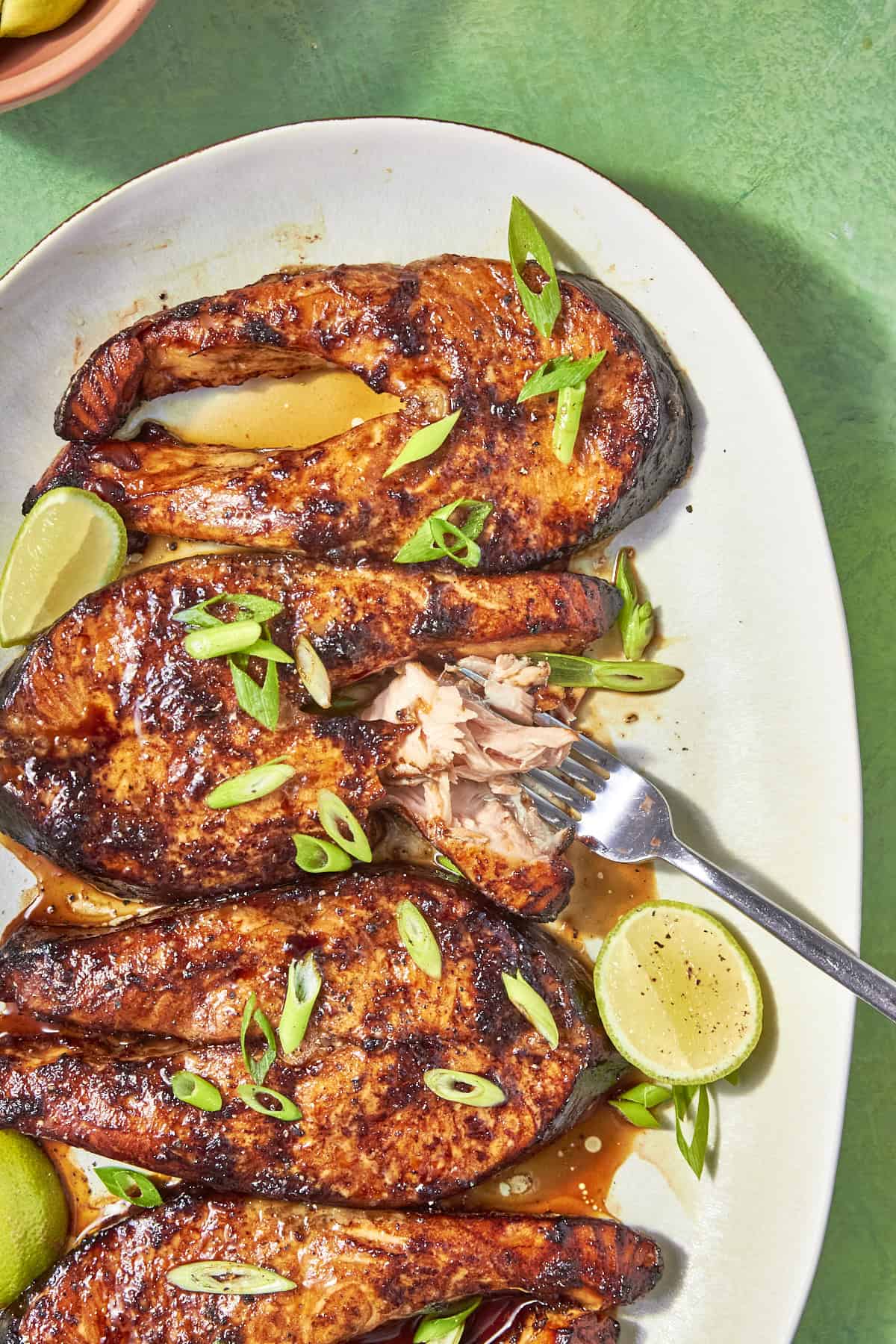 Oven Baked Salmon Steaks with a caramelized glaze, garnished with sliced green onions and lime halves, served on a white oval plate. A fork has flaked one piece of salmon. The background is a green surface.