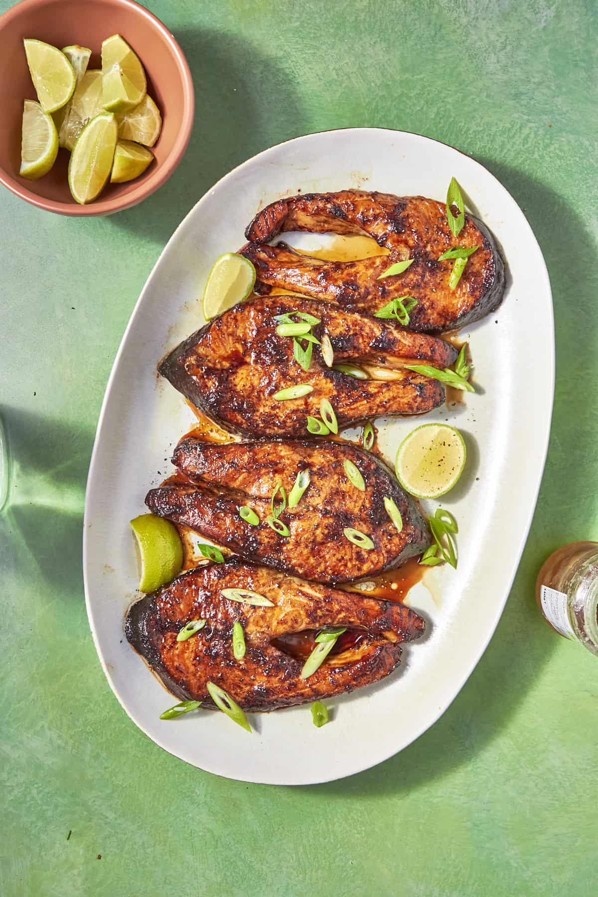An oval white plate with four pieces of oven baked salmon steaks garnished with sliced green onions and lime wedges, set on a green surface next to a small bowl of lime wedges and a glass bottle.