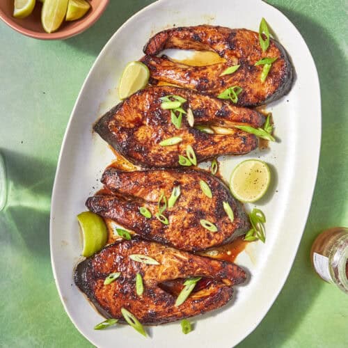 A white oval plate holds four spiced, oven baked salmon steaks garnished with sliced green onions and lime wedges, set on a green surface. A bowl of lime wedges is nearby.