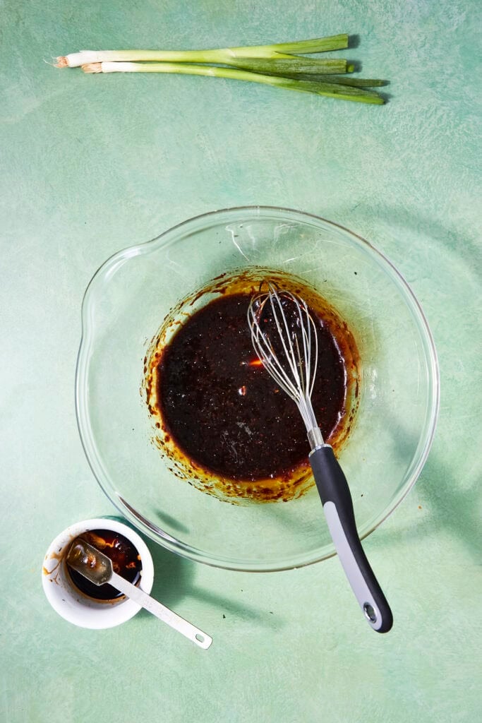 A glass bowl with dark sauce and a whisk sits on a green countertop—perfect for marinating Oven Baked Salmon Steaks. Nearby are a small bowl with sauce, a spoon, and whole green onions at the top of the image.