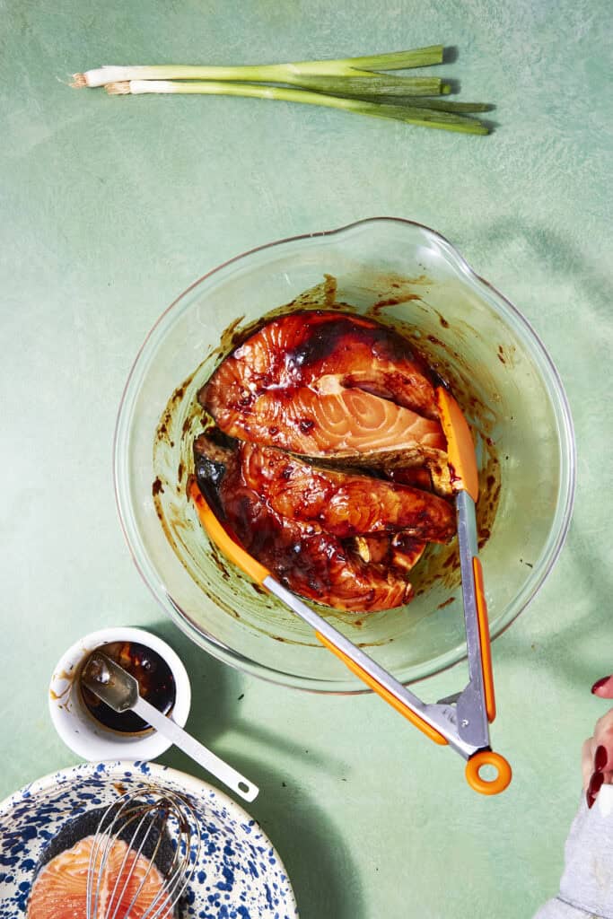 A glass bowl with raw salmon fillets marinating in a dark sauce for Oven Baked Salmon Steaks, held by orange-handled tongs. Nearby are green onions, a whisk, a small bowl of sauce, and a plate with more salmon.
