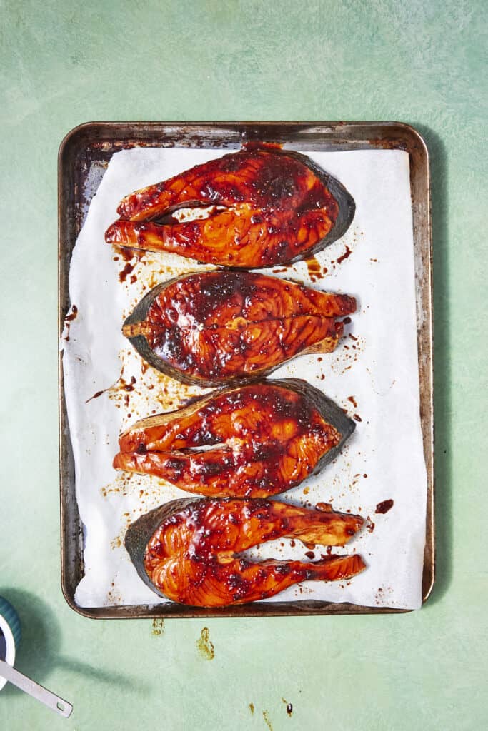 Four Oven Baked Salmon Steaks are arranged in a single row on a parchment-lined baking sheet, topped with a shiny sauce and sprinkled with sesame seeds, set against a pale green background.