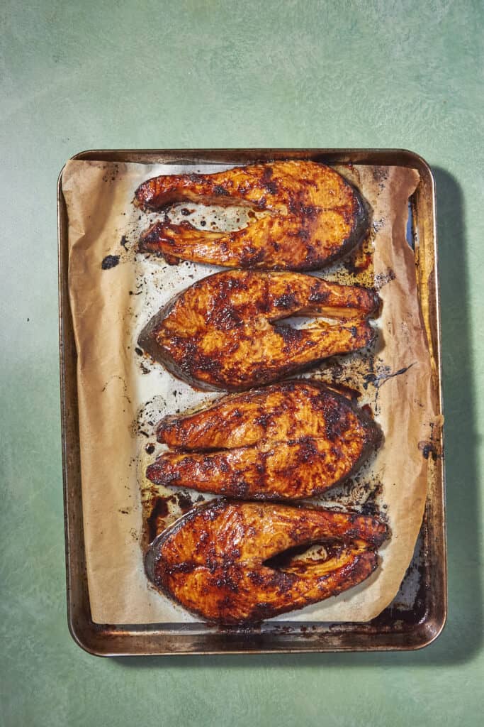 Four Oven Baked Salmon Steaks with a reddish seasoning are arranged in a row on a parchment-lined baking sheet atop a green surface. The fish is beautifully browned and slightly charred around the edges.