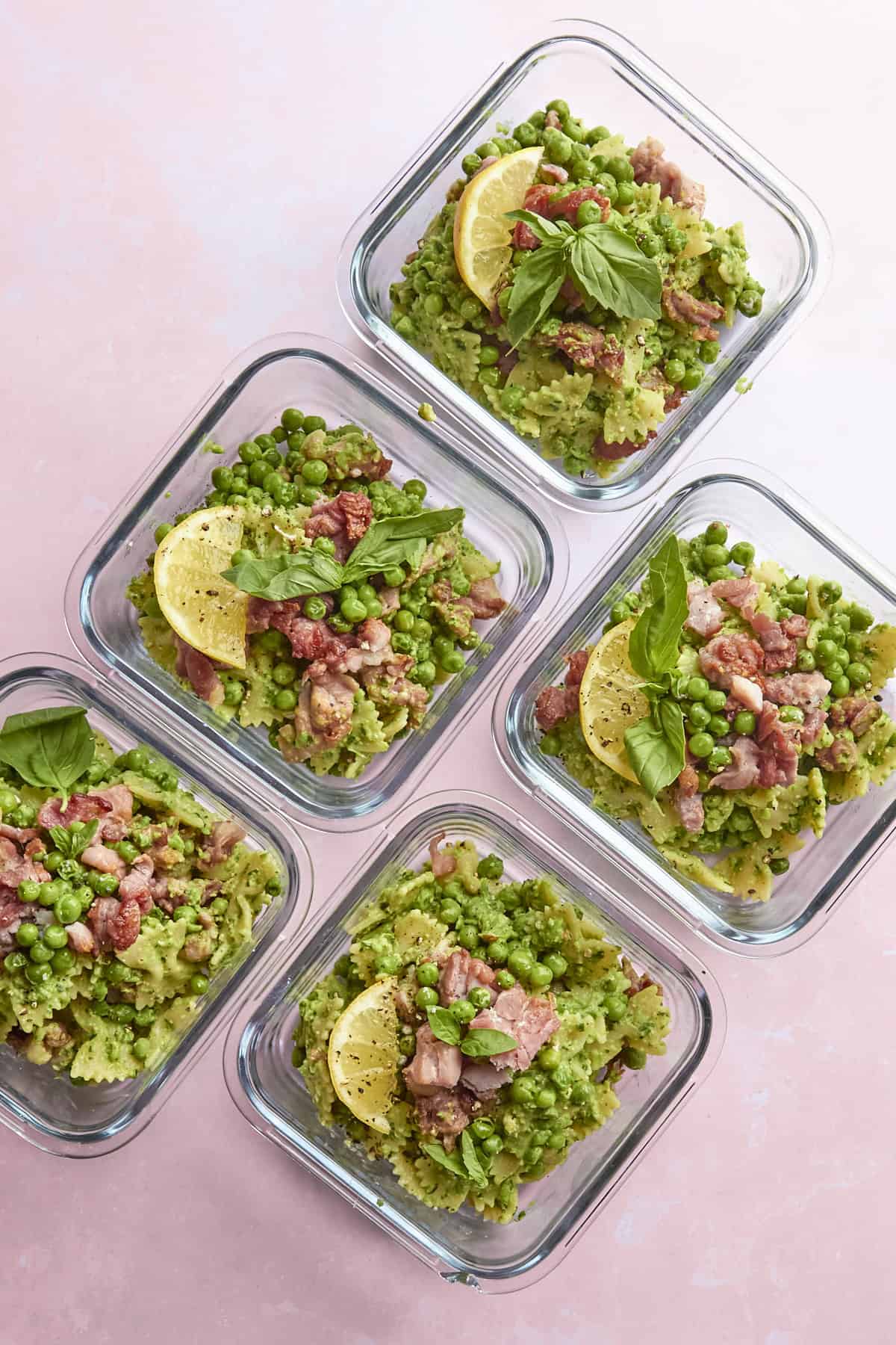 Five glass containers filled with pasta mixed with green peas, pieces of bacon, roasted corn, fresh basil leaves, and lemon slices, arranged on a pink background.