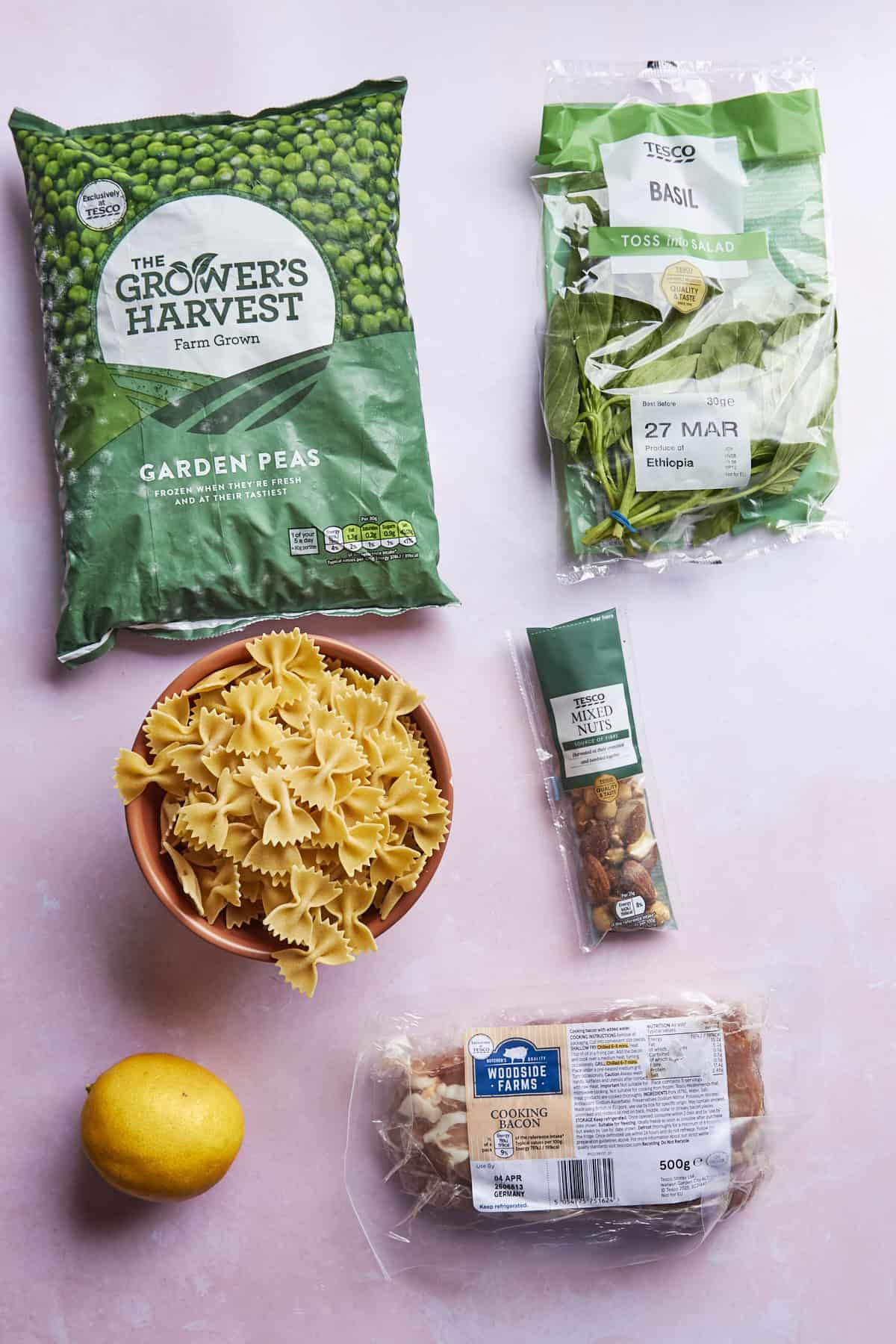A flat lay of ingredients on a pink surface: a bag of garden peas, roasted corn, a bag of basil, a bowl of farfalle pasta, a lemon, a pack of chopped walnuts, and a loaf of white bread.