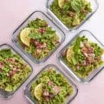 Five glass containers filled with bow tie pasta, green peas, diced meat, roasted corn, and garnished with lemon slices and basil leaves, arranged on a light pink surface.