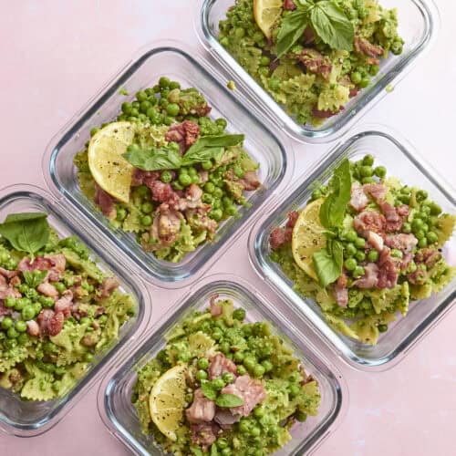 Five glass containers filled with bow tie pasta, green peas, diced meat, roasted corn, and garnished with lemon slices and basil leaves, arranged on a light pink surface.