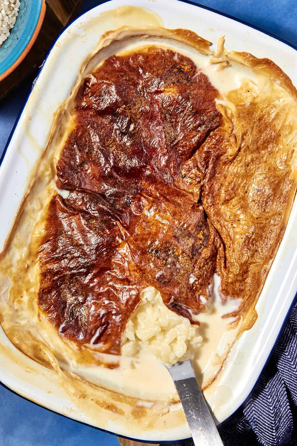 A baking dish filled with creamy rice pudding is topped with a golden, caramelized skin. A serving of rice pudding has been scooped out, revealing the creamy rice beneath the browned surface. A metal spoon rests inside the dish.