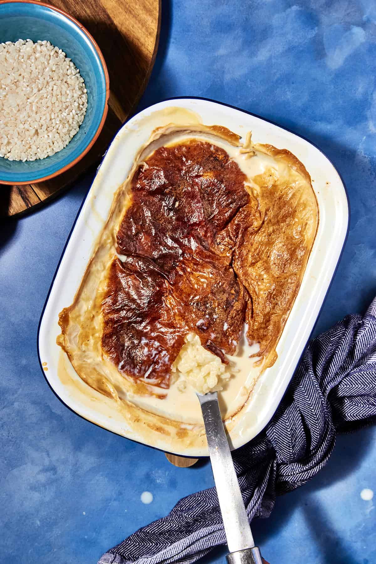 A baked rice pudding with a golden brown top sits in a white dish on a blue surface, a serving spoon inside. Beside the creamy rice pudding, a bowl of uncooked rice and a striped cloth napkin are visible.