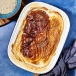 A classic rice pudding is baked in a white rectangular dish with a golden, caramelized top, set on a blue surface next to a bowl of uncooked rice and a striped cloth napkin.