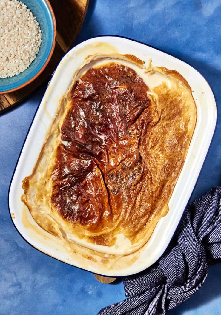 A classic rice pudding is baked in a white rectangular dish with a golden, caramelized top, set on a blue surface next to a bowl of uncooked rice and a striped cloth napkin.