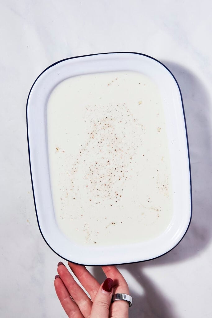 A hand with red nail polish holds a white enamel dish filled with creamy rice pudding, sprinkled with ground spices, on a light marble surface.