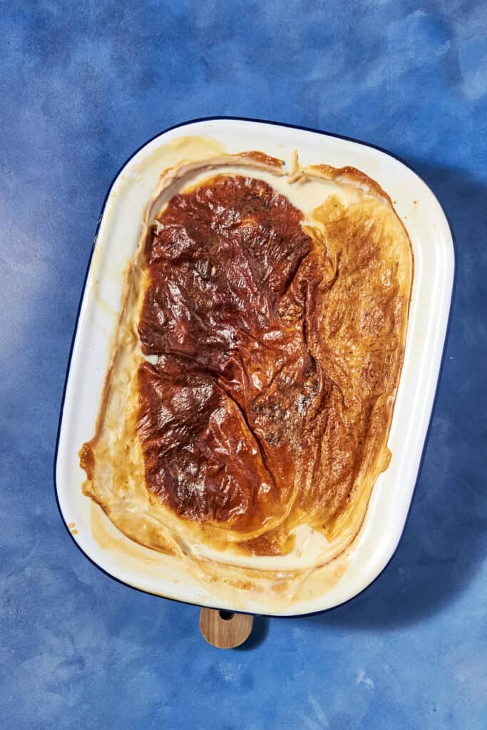 A rectangular baking dish filled with golden-brown rice pudding sits on a blue textured surface. The baked custard dessert has a slightly wrinkled, caramelized top layer.