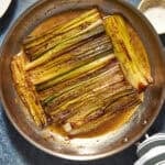 Leeks sautéed and caramelized in a skillet with a savory sauce and roasted corn, sprinkled with spices, on a blue surface near a small bowl of grated cheese and a striped cloth.