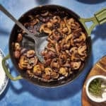 A skillet filled with savory sauteed mushrooms sits on a blue surface, surrounded by a metal spatula, a wooden board with a small bowl of herbs, and partial views of decorative plates.
