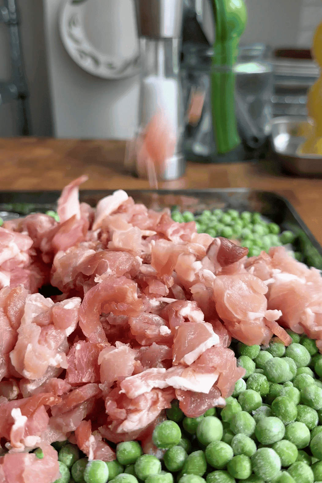 A close-up of diced raw bacon pieces piled on top of a tray filled with green peas and roasted corn, with a blurred kitchen background.