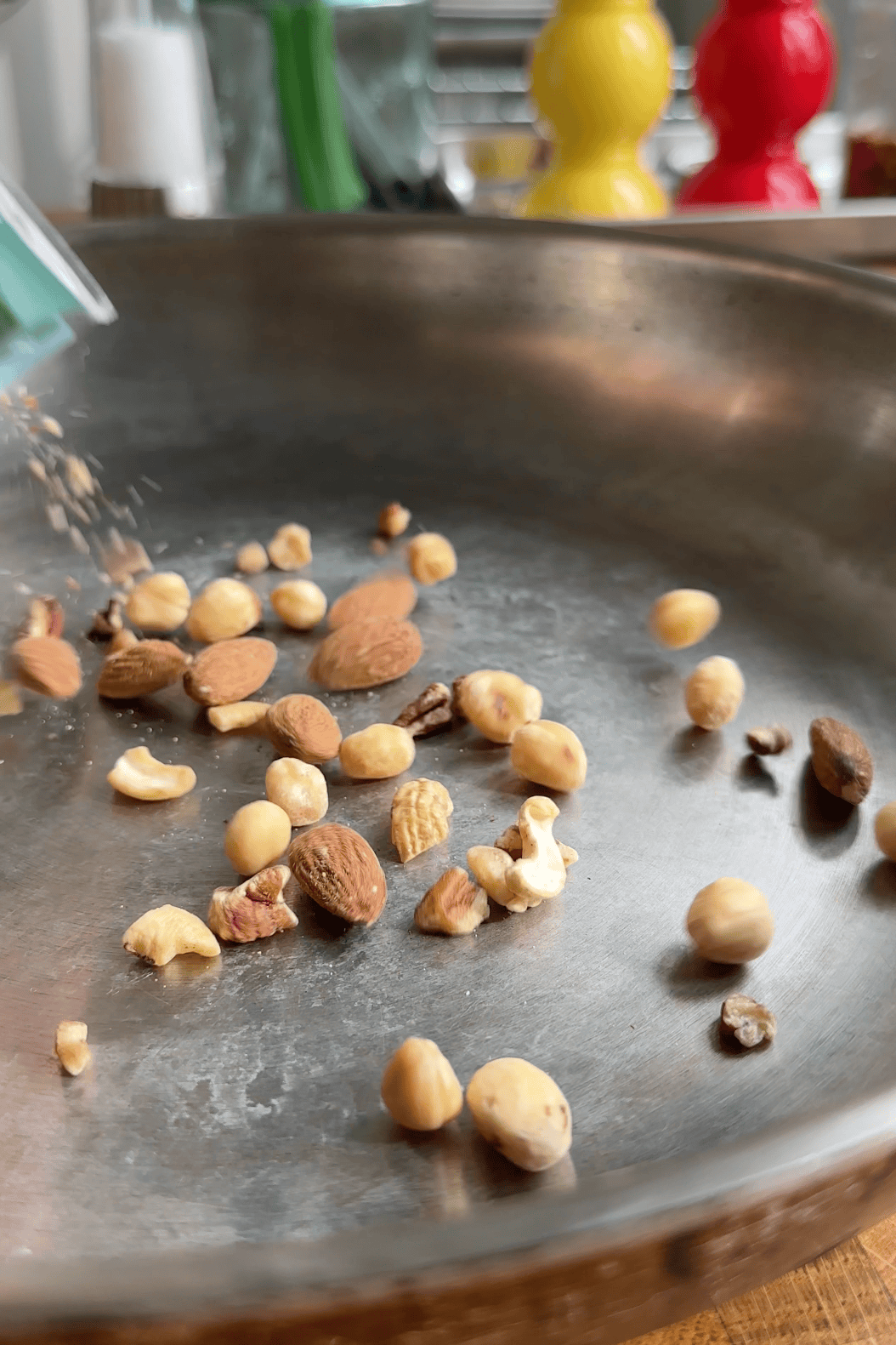 A close-up of assorted raw nuts, including almonds, hazelnuts, cashews, and roasted corn, being poured into a stainless steel pan. The background features blurred yellow and red items, likely kitchenware.