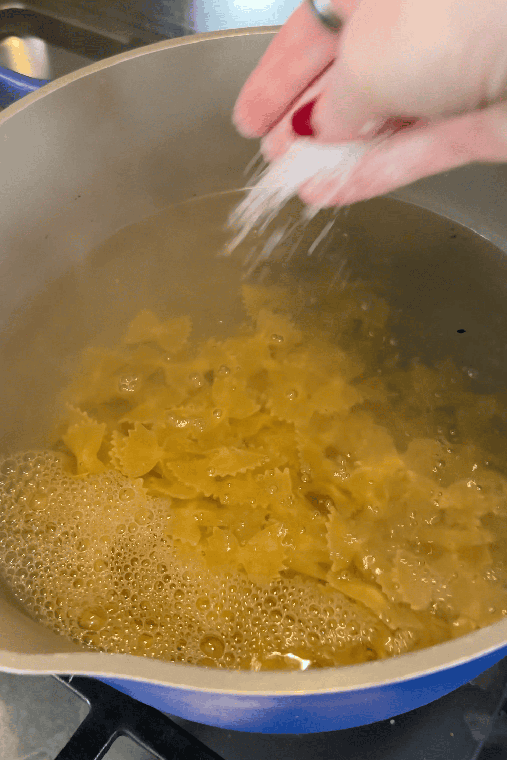 A hand sprinkles salt into a pot of boiling water with farfalle pasta cooking inside on a stovetop, ready to be paired with roasted corn for a delicious meal.