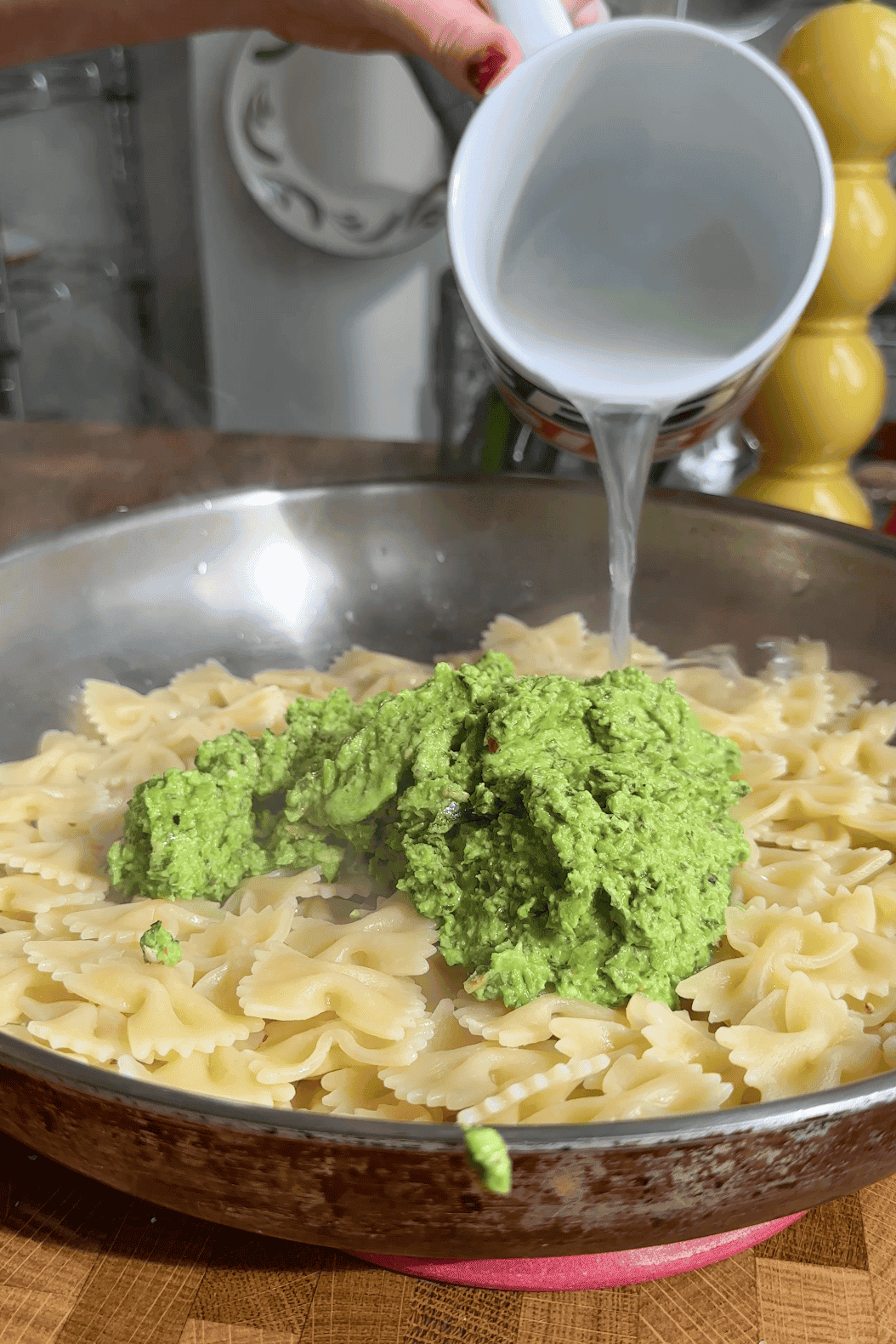 A close-up of cooked bow-tie pasta in a pan with a large spoonful of green pesto and roasted corn on top. A hand pours liquid, likely pasta water, from a small white cup into the pan.