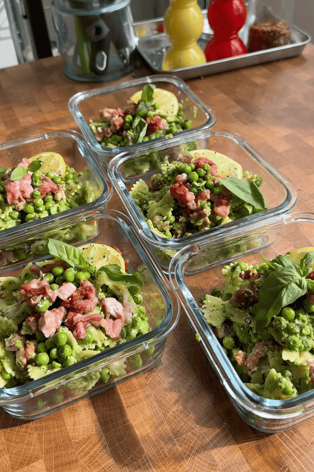 Five glass containers on a wooden counter, each filled with pasta, green peas, roasted corn, bacon pieces, fresh basil leaves, and a slice of lemon, ready for meal prep. Kitchen items are blurred in the background.