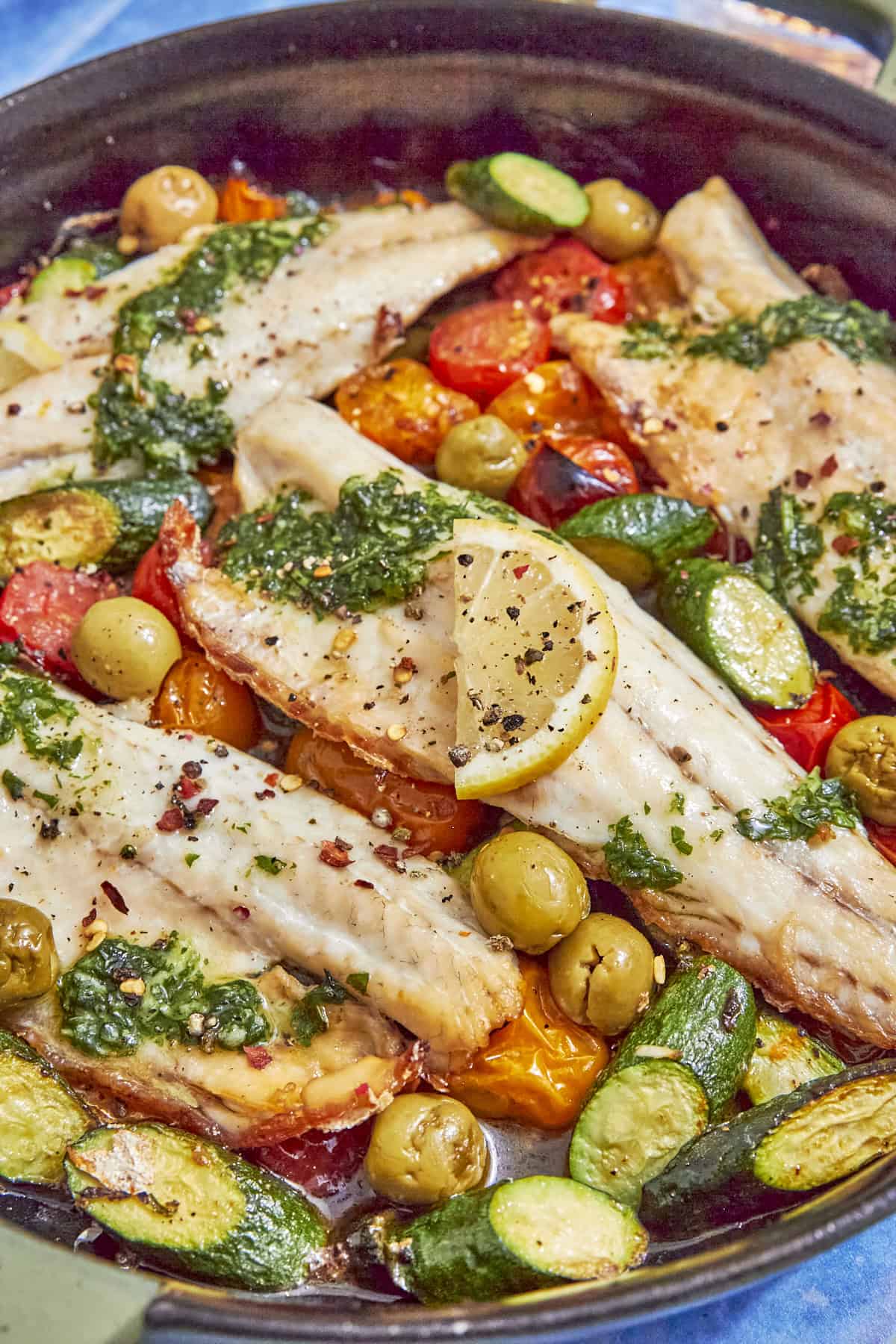 A skillet filled with baked sea bass fillets topped with green herb sauce, lemon slices, cherry tomatoes, olives, and sliced zucchini. This sea bass recipe appears colorful and is beautifully garnished with spices.