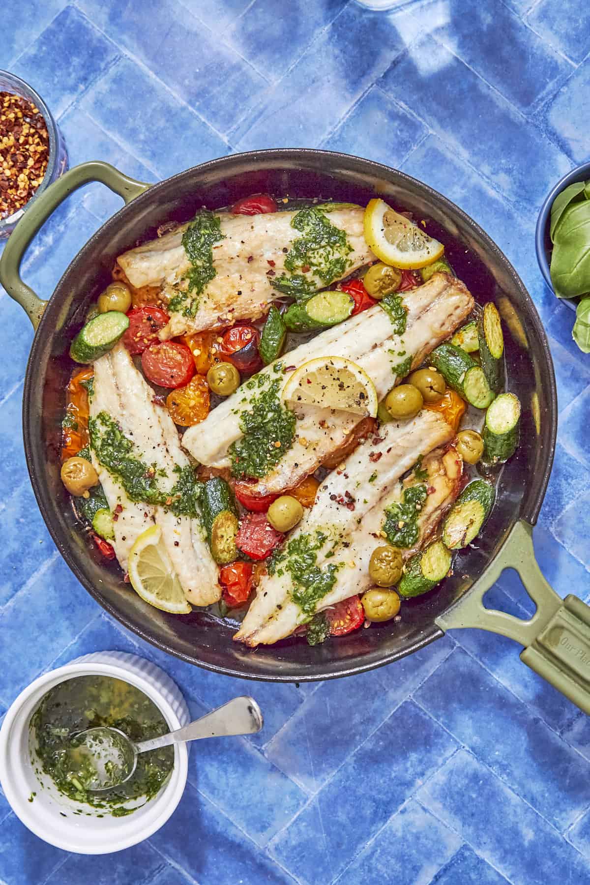 A green pan filled with baked sea bass fillets, cherry tomatoes, sliced zucchini, olives, lemon slices, and green herb sauce on a blue tile surface, with a small bowl of sauce and basil leaves nearby—an inviting sea bass recipe.